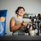 Eine Frau schäumt Milch auf an einer Rocket Apartamento. Im Vordergrund der Kaffee Mocino Salvatore