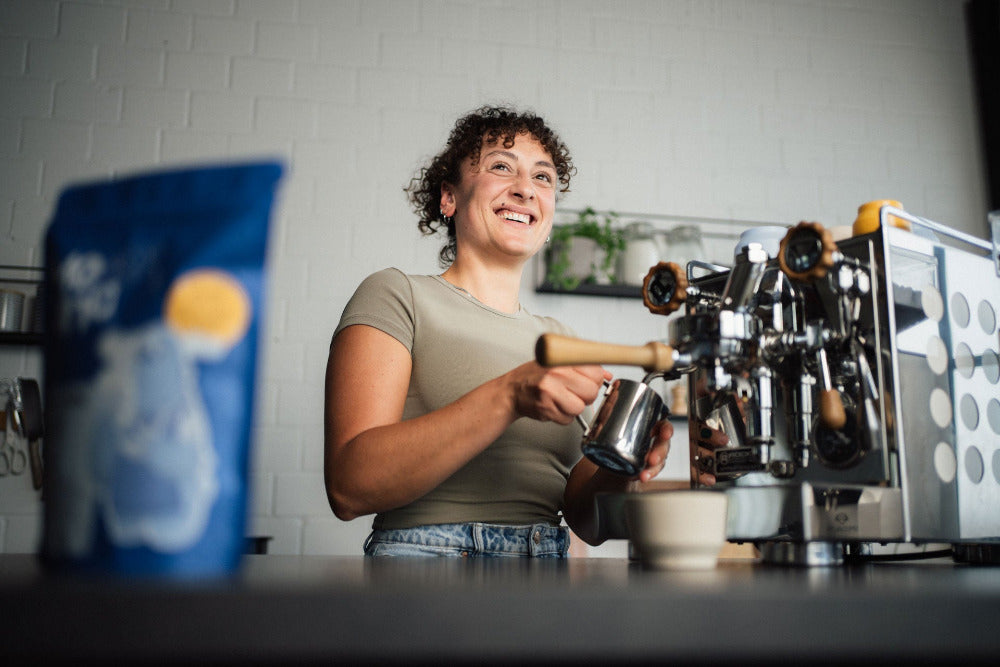 Eine Frau schäumt Milch auf an einer Rocket Apartamento. Im Vordergrund der Kaffee Mocino Salvatore